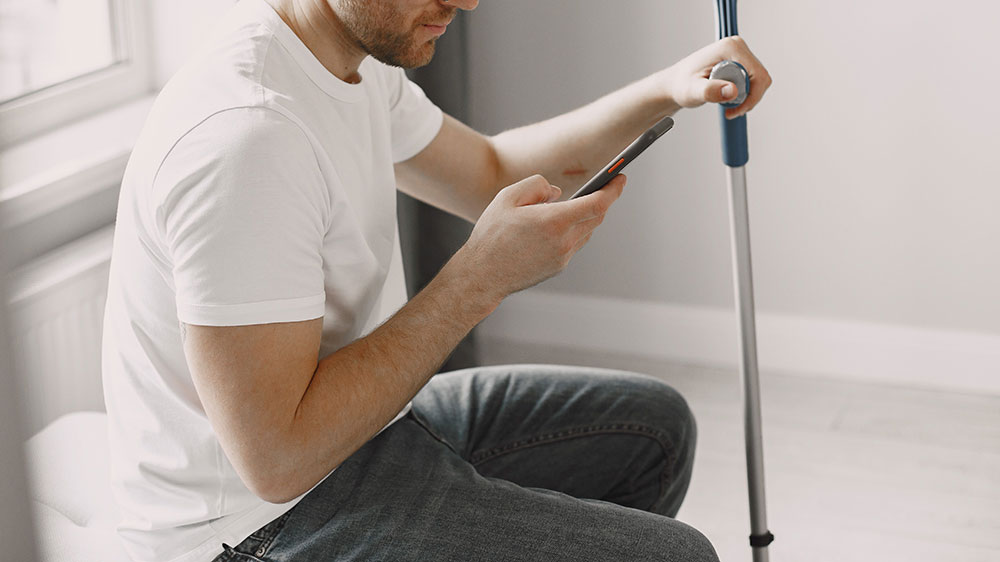 Adult man with eyeglasses using a smartphone indoors, with a crutch beside him.